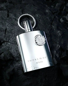 The image shows a silver perfume bottle from the "SUPREMACY" line by AFNAN. The bottle is placed on a textured, dark stone background that contrasts with its shiny metallic finish. It has a modern, sleek design with a reflective surface, and is accented with a circular metallic loop atop its cap. An intricate emblem is embossed on the bottle, adding to the luxurious look.   Water droplets are visible on the bottle, suggesting a fresh appeal and highlighting the bottle's metallic sheen. 