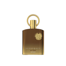The image shows a luxurious perfume bottle with a sleek, modern design. The bottle appears to be made of a glossy, brown-gold glass with a golden cap that has a circular handle on top, giving it a unique and elegant look. The front of the bottle features a prominent emblem, likely a brand logo, embossed in gold which adds to the opulent aesthetic. Below the emblem, the words "SUPREMACY in Oud AFNAN" are printed in capital letters, indicating the fragrance name and brand. 