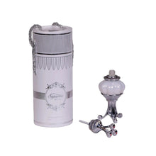 The image displays two items related to fragrance products on a white background. To the left is a cylindrical container adorned with a decorative silver chain, labeled "Supreme MUSK" with additional text below stating "Concentrated Perfume Oil 20ml." The container has a white label with an intricate, decorative border and a classic ornate emblem in the center.  The bottle's top has a textured design with a tapered middle and flared ends, characteristic of vintage perfume dabber.
