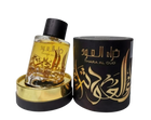 A clear glass perfume bottle with a yellow-amber liquid inside is displayed next to a cylindrical black packaging with gold lettering. The bottle has a dark cap and a gold label with Arabic calligraphy that reads "THARA AL OUD." The packaging repeats the name "THARA AL OUD" in gold Arabic calligraphy and has intricate gold decorative elements. Both the bottle and packaging sit atop a golden base, and the background is neutral, focusing attention on the items.