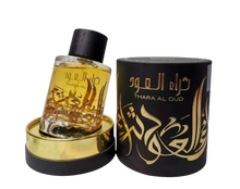 A clear glass perfume bottle with a yellow-amber liquid inside is displayed next to a cylindrical black packaging with gold lettering. The bottle has a dark cap and a gold label with Arabic calligraphy that reads "THARA AL OUD." The packaging repeats the name "THARA AL OUD" in gold Arabic calligraphy and has intricate gold decorative elements. Both the bottle and packaging sit atop a golden base, and the background is neutral, focusing attention on the items.