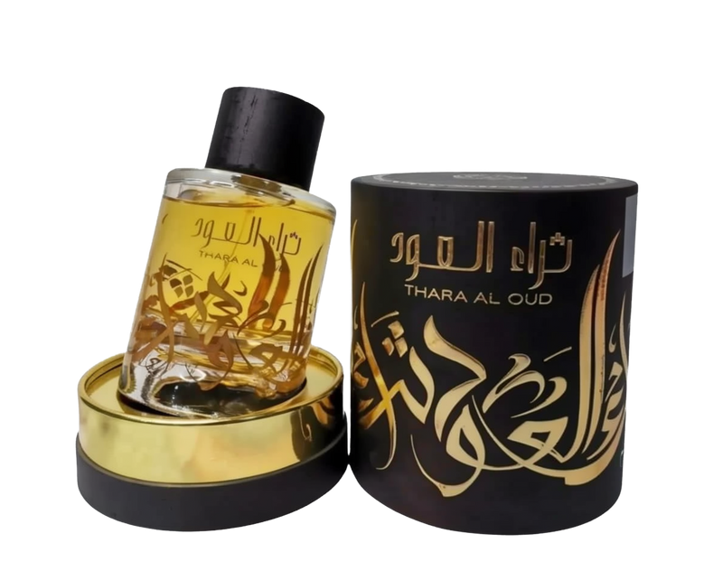 A clear glass perfume bottle with a yellow-amber liquid inside is displayed next to a cylindrical black packaging with gold lettering. The bottle has a dark cap and a gold label with Arabic calligraphy that reads "THARA AL OUD." The packaging repeats the name "THARA AL OUD" in gold Arabic calligraphy and has intricate gold decorative elements. Both the bottle and packaging sit atop a golden base, and the background is neutral, focusing attention on the items.