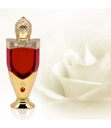 The image features an ornate perfume bottle prominently displayed in the foreground. The bottle has a vivid red center, capped with an elaborate gold lid that has a diamond-like pattern, topped with a decorative finial resembling a gemstone. A matching golden base supports the bottle, with a red gemstone inset just above the base. The background is softly focused, featuring what appears to be a white rose in the right corner, adding a sense of luxury and suggesting the fragrance might have floral notes.