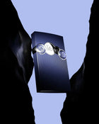 The image depicts a navy blue rectangular box with a ribbed texture, situated between what appears to be two dark, jagged rock formations against a blue sky background. The box is embellished with three circular, silver-colored emblems placed in a horizontal line, with the central emblem featuring the word "Tribute" in an elegant script. The lighting emphasizes the box and the metallic sheen of the emblems, creating a dramatic and luxurious presentation.