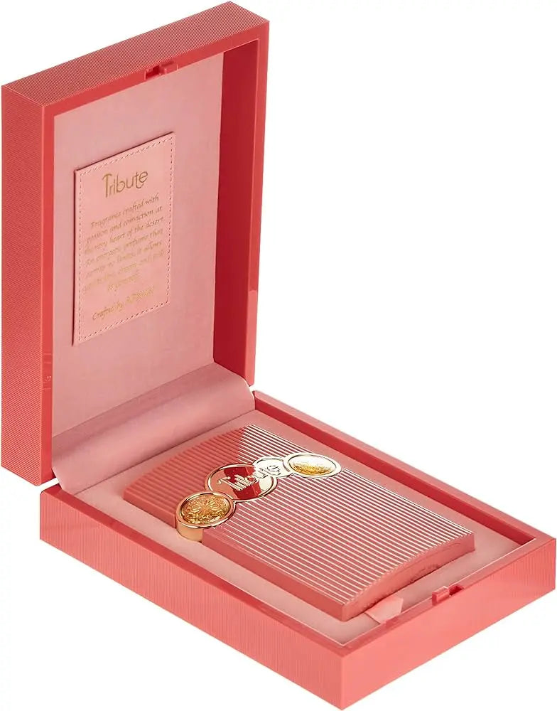 The image features an open pink box on a white background. The interior of the box is a lighter shade of pink, with the left side displaying a framed panel that reads "Tribute" at the top in an elegant script, followed by descriptive text. This text appears to be a narrative or description related to the product, bordered by a thin golden frame. On the right side of the box, there is a recessed area holding a ribbed, pink rectangular object with three circular golden emblems attached to it. 