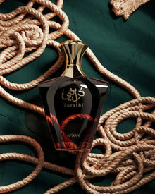 The image features a luxurious perfume bottle placed on a textured dark teal background intertwined with thick, twisted ropes that appear to be made of a light brown fiber. The perfume bottle has a sleek, dark exterior with a reflective surface, highlighting a golden Arabic calligraphy emblem in the center, above which the word "Turathi" is written in a stylized font. Below the emblem is the word "AFNAN." The cap of the bottle is gold and faceted, complementing the elegant design. 