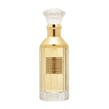 The image is of a perfume bottle:  The bottle has a rectangular shape with clear glass, revealing the golden-colored liquid perfume inside. It features a gold-colored neck and a white, bristle-like applicator that resembles a traditional shaving brush. The front of the bottle has a simple, elegant label with "VELVET OUD" in black letters and Arabic script below it.