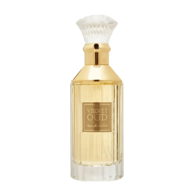 The image is of a perfume bottle:  The bottle has a rectangular shape with clear glass, revealing the golden-colored liquid perfume inside. It features a gold-colored neck and a white, bristle-like applicator that resembles a traditional shaving brush. The front of the bottle has a simple, elegant label with "VELVET OUD" in black letters and Arabic script below it.