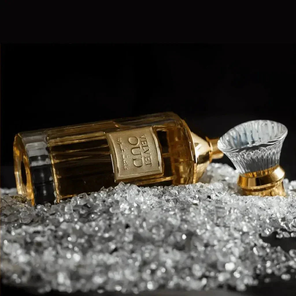 The image depicts a perfume bottle laid on a bed of crystals:  The bottle is rectangular with clear glass, filled with a golden-colored perfume, suggesting a warm scent. It has a distinctive white, bristle-like applicator that looks similar to a traditional shaving brush, with a gold band around its base. The cap is off and placed beside the bottle, showing the applicator.
