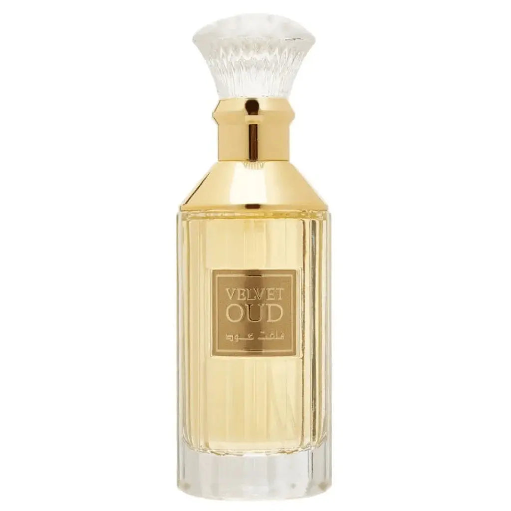 The image is of a perfume bottle:  The bottle has a rectangular shape with clear glass, revealing the golden-colored liquid perfume inside. It features a gold-colored neck and a white, bristle-like applicator that resembles a traditional shaving brush. The front of the bottle has a simple, elegant label with "VELVET OUD" in black letters and Arabic script below it.