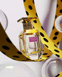 The image shows a perfume bottle named "VIOLET BOUQUET" by AFNAN. The bottle is made of clear glass with a hexagonal faceted base and a polished gold cap. It features a braided gold band around the neck and a purple label with the perfume name in white font. The background is creatively designed with abstract elements including a yellow ribbon with black polka dots and a white sphere, creating a playful and artistic setting for the perfume. 