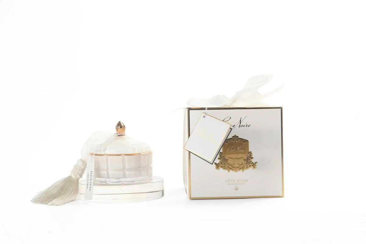 The image features an elegant Cote Noire product display on a white background. On the left, a decorative candle in a translucent white glass container with a golden knob on top is adorned with a gray tassel, adding a touch of sophistication. Next to the candle is a luxurious white gift box with gold trim, tied with a white ribbon. A gift tag attached to the box reads "Cote Noire" in stylish script.