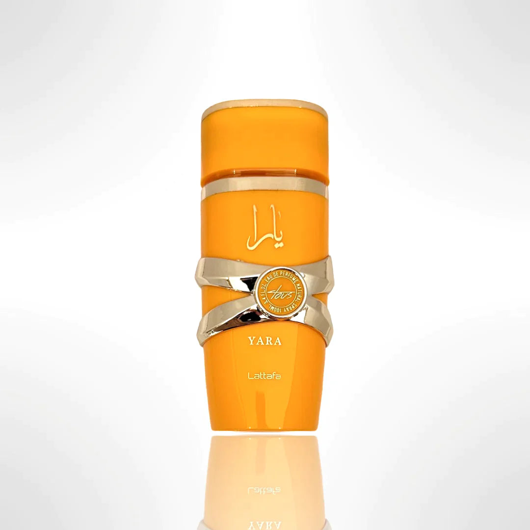 This image features a bright orange cylindrical perfume bottle with the name "YARA" from the brand Lattafa. The design is distinguished by a gold ribbon-like band wrapped around the center, with a circular emblem featuring text that may denote "EAU DE PARFUM NATURAL SPRAY". The Arabic script for "YARA" is placed above the gold band. The bottle has a glossy finish and is presented against a gradient background that fades from white at the top to a light reflection of the bottle at the base.