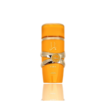 This image features a bright orange cylindrical perfume bottle with the name "YARA" from the brand Lattafa. The design is distinguished by a gold ribbon-like band wrapped around the center, with a circular emblem featuring text that may denote "EAU DE PARFUM NATURAL SPRAY". The Arabic script for "YARA" is placed above the gold band. The bottle has a glossy finish and is presented against a gradient background that fades from white at the top to a light reflection of the bottle at the base.