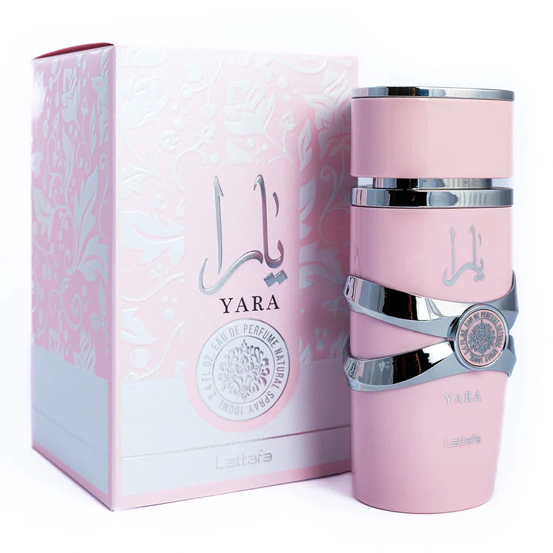 This image features the perfume "YARA" by Lattafa, displayed next to its packaging. The cylindrical bottle is pale pink with silver bands, mirroring the previous description. The packaging is a square box with a matching pale pink color and a subtle floral pattern embossed on it, adding a delicate texture. The perfume name "YARA" is prominently displayed in a darker pink shade with the Arabic script above and the phrase "EAU DE PERFUME NATURAL" in a circular silver emblem. 