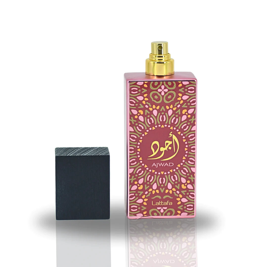 Image of a tall pink perfume bottle with a gold spray nozzle, placed upright next to its black textured cap which is lying on its side. The bottle has a decorative pattern in a lighter pink hue with a central circular label containing gold Arabic calligraphy and the word 'AJWAD'. Below the pattern, 'Lattafa' is written in small gold letters. The bottle is reflected on a glossy surface below, indicating a polished presentation.