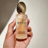 The image shows a hand holding a bottle of hair mist. The bottle has a frosted glass appearance with a gradient of color, possibly indicating the scent or type of the hair mist. It has a gold cap that appears textured, resembling a classic perfume bottle design. Arabic script is visible on the front label, alongside the English translation which reads "BADEE AL OUD" and "FRESH HAIR MIST.