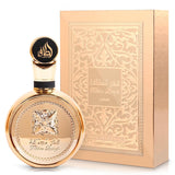 An image of a golden perfume bottle with a round body and a large, circular black cap with gold Arabic calligraphy and the name 'Lattafa'. The central part of the bottle displays an embossed flower design, with the name 'Fakhar Lattafa' inscribed below. Next to the bottle is its packaging, a golden box with intricate white arabesque patterns and a central rectangle that reads 'Fakhar Lattafa' in both Arabic and English.