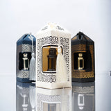 An image of three ornate perfume bottles, each enclosed in a decorative lantern-like casing with intricate cut-out designs and tassels. The central casing is white with black patterns and a tassel, showcasing a clear bottle with dark liquid inside. Flanking it are two casings, one black with silver patterns and the other black with gold patterns, each containing similar bottles. The background is reflective, creating mirror images of the casings and bottles.