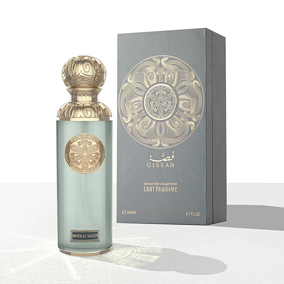 The image features a perfume bottle and its packaging from the "Gissah" Signature Collection, described as a light fragrance. The bottle is tall and cylindrical with a frosted light teal glass body, adorned with ornate gold medallions. It is capped with a gold, intricately designed spherical lid. Beside the bottle is the box, which is gray with a textured appearance and a large gold mandala-like design on the front, with the name "Gissah" printed at the top.