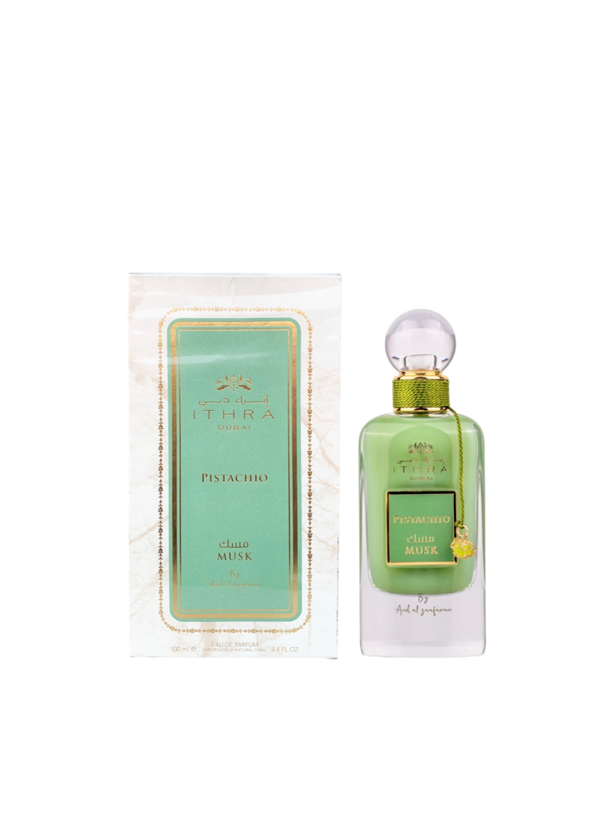 This image shows a bottle of Ithra Dubai Pistachio Musk perfume next to its packaging. The perfume bottle has a green hue with a clear spherical cap and a gold-threaded neck, adorned with a small gold charm and green string. The box has a marble-like design with gold and green accents, reflecting the luxurious and elegant branding. It features the brand name Ithra Dubai and the fragrance name Pistachio Musk, with additional text in Arabic, indicating a 100ml eau de parfum.