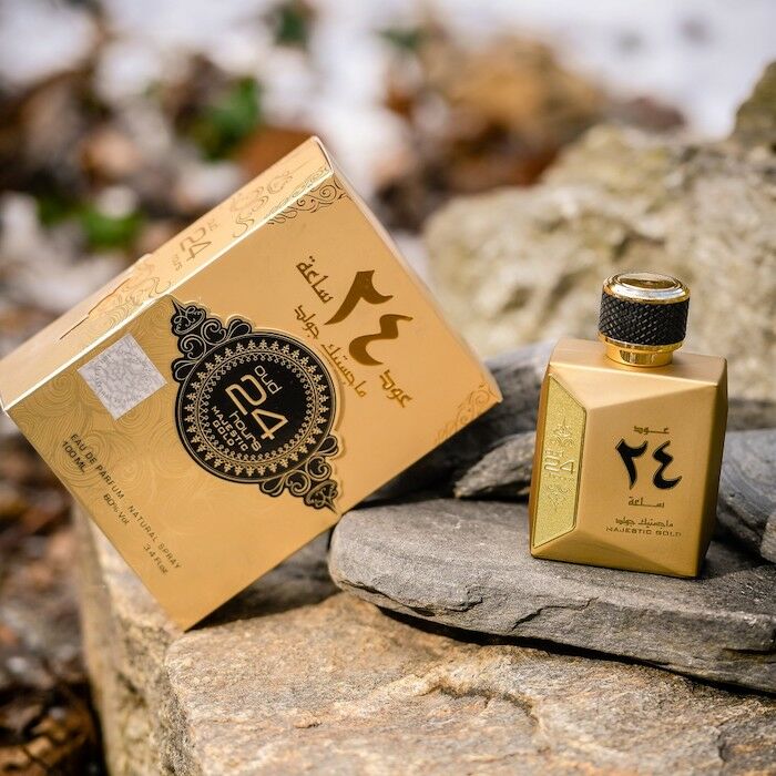 The image features a small, golden perfume bottle with a textured black cap, placed on a stone surface. Beside the bottle is its box, which has a beige background and is adorned with intricate black and gold designs, as well as Arabic calligraphy and the phrase "MAJESTIC GOLD" in English. The emblem of the brand is prominent in the center of the box. The natural outdoor setting with stones and dry leaves in the background gives the product a rustic and elegant presentation.